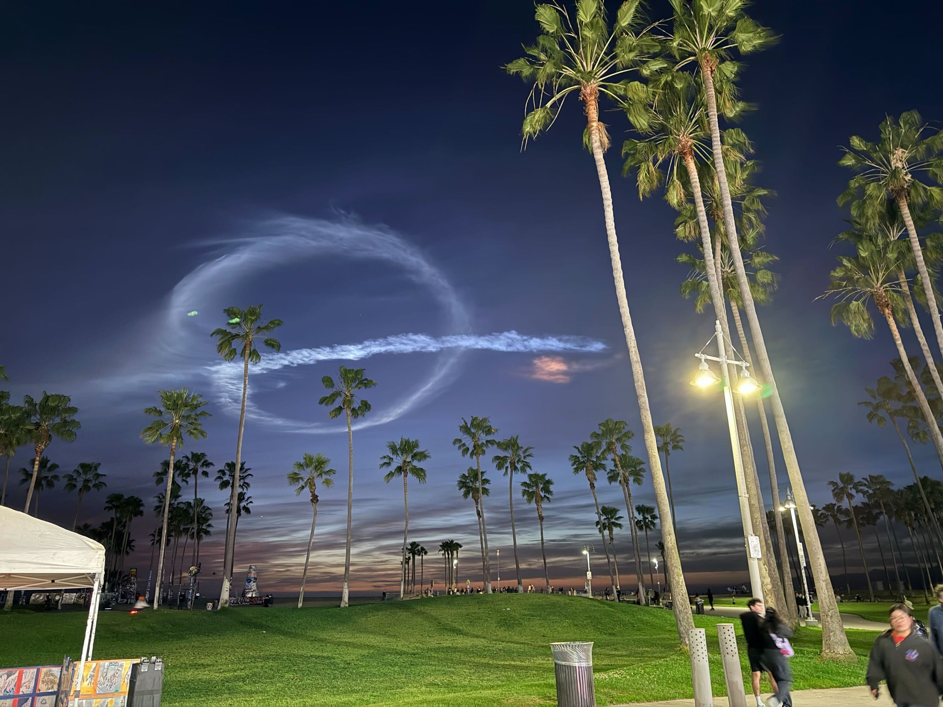 SpaceX rocket trails forming spiral patterns in the twilight sky over Venice Beach, with palm trees silhouetted against the colorful sunset